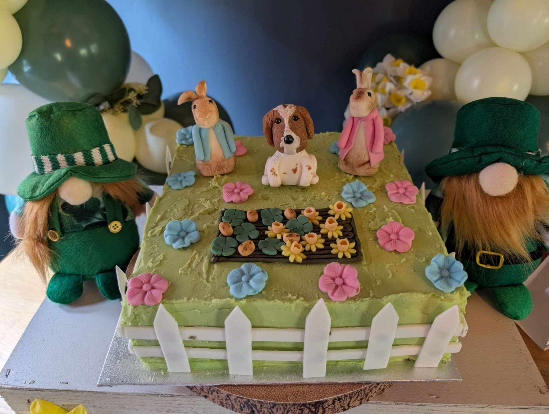 Green garden-themed square cake with fondant rabbits, a dog, flowers and a picket fence