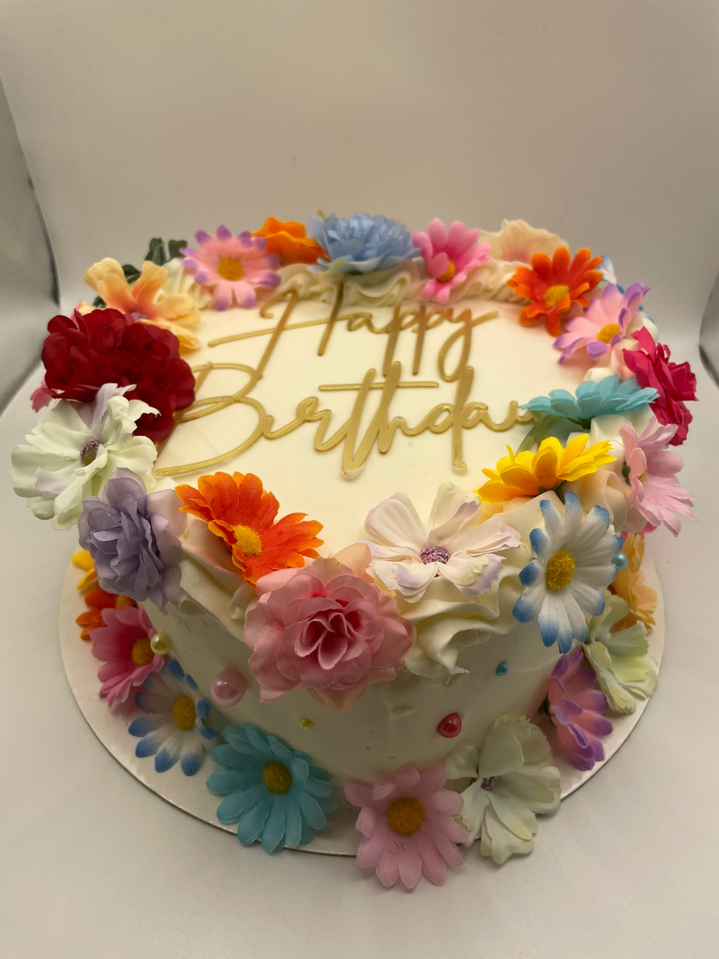 Buttercream birthday cake surrounded by a wreath of colourful flowers with a gold Happy Birthday topper